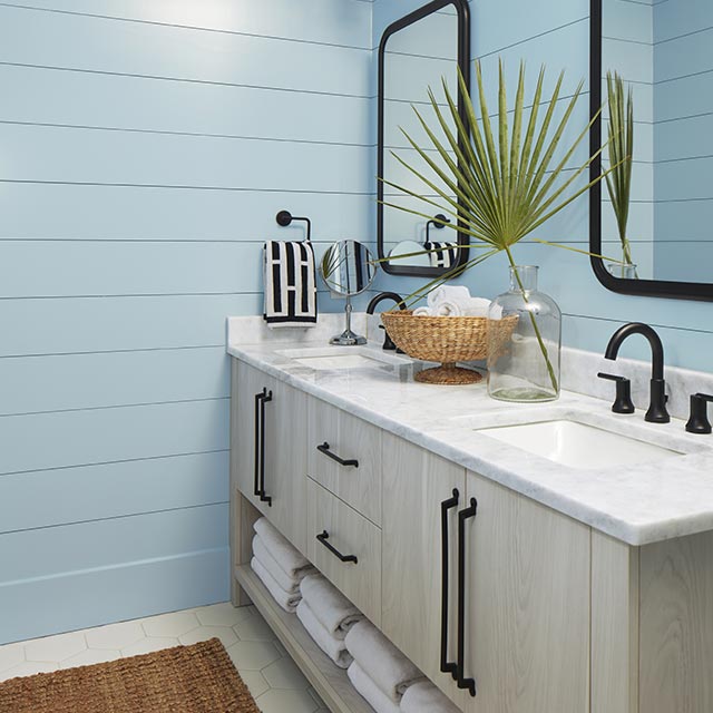 A bathroom with pretty blue-painted shiplap walls, a white ceiling, a blonde wood double vanity with a marble top and black fixtures, and two mirrors. A bathroom with pretty blue-painted shiplap walls, a white ceiling, a blonde wood double vanity with a marble top and black fixtures, and two mirrors.