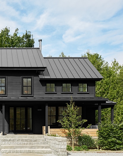 Gray stone masonry leads up to a modern home with black siding and shed-style aluminum black roofing. Gray stone masonry leads up to a modern home with black siding and shed-style aluminum black roofing.