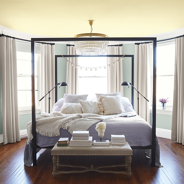 Rounded bedroom walls painted a soft blue-gray, a pale yellow ceiling, white trim, and three windows surround a comfy black four-poster bed. Rounded bedroom walls painted a soft blue-gray, a pale yellow ceiling, white trim, and three windows surround a comfy black four-poster bed.