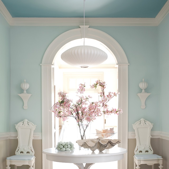 A spacious hallway with light blue painted upper walls, neutral lower walls, and a blue ceiling features a white table and chairs, and a white painted arch and trim throughout.   A spacious hallway with light blue painted upper walls, neutral lower walls, and a blue ceiling features a white table and chairs, and a white painted arch and trim throughout.