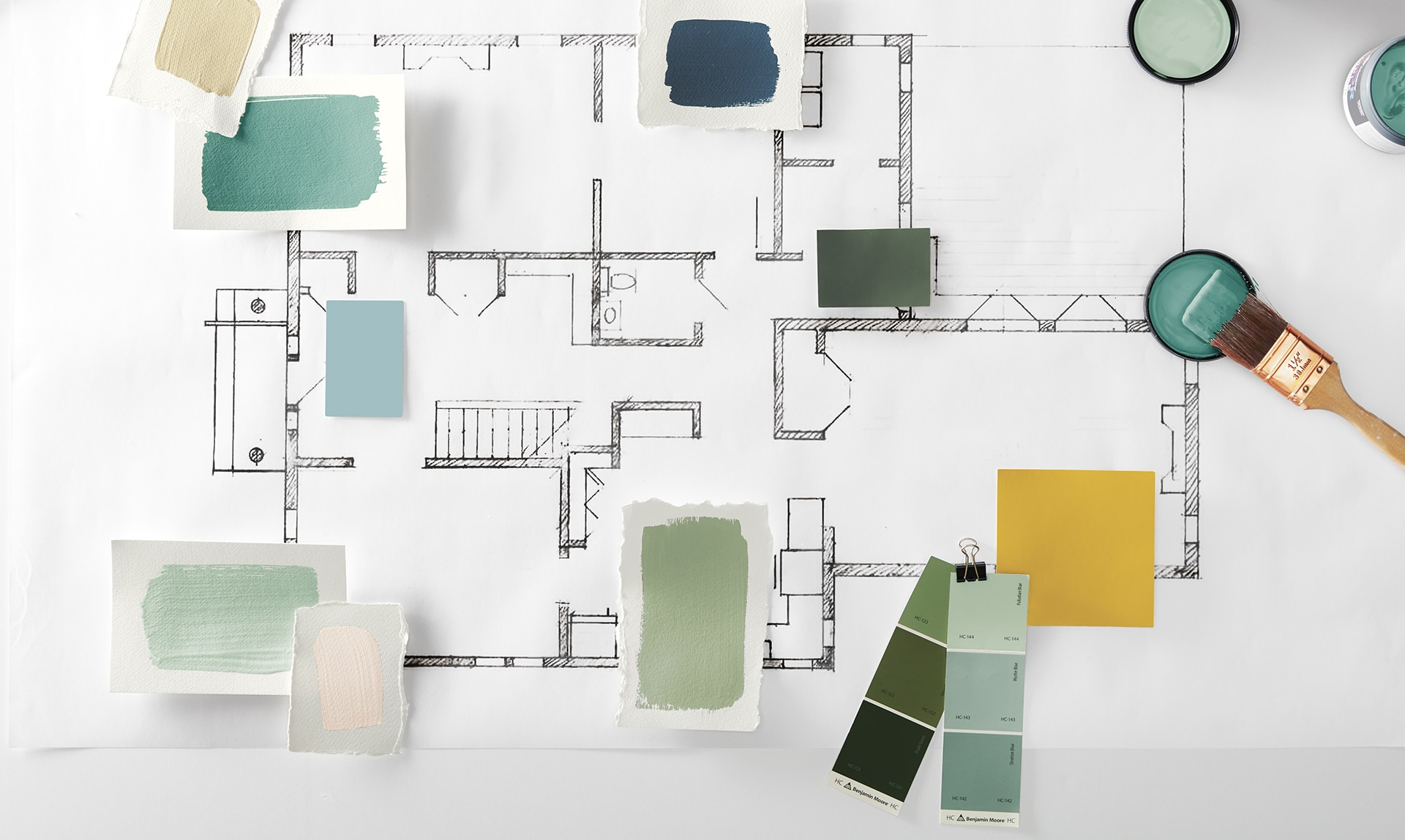 A floor plan with shades of green and blue paint samples placed on top, along with multiple open cans of green paint and a paint brush.