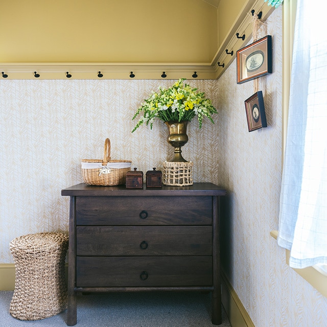 A corner space with dark tan painted upper walls, trim, and ceiling, tan printed wallpaper, and a wooden bureau with wicker decor. A corner space with dark tan painted upper walls, trim, and ceiling, tan printed wallpaper, and a wooden bureau with wicker decor.