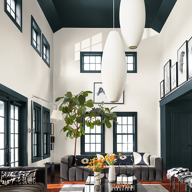 A sunlit living room with two-story white painted walls; a deep green vaulted ceiling, trim, and doors; large oval pendant lights; and neutral modern decor. A sunlit living room with two-story white painted walls; a deep green vaulted ceiling, trim, and doors; large oval pendant lights; and neutral modern decor.