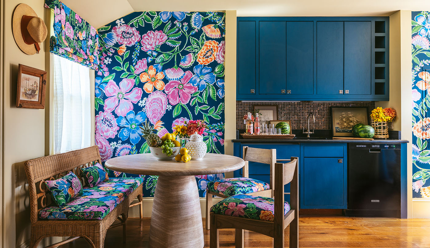 A cheerful dining and kitchen area with vibrant floral wallpaper and matching floral seat cushions, dark tan painted walls and ceiling, and dark blue cabinets.