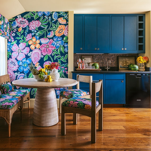 A cheerful dining and kitchen area with vibrant floral wallpaper and matching floral seat cushions, dark tan painted walls and ceiling, and dark blue cabinets.