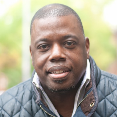A headshot of an African American male wearing a white button down and navy blue barbour jacket.