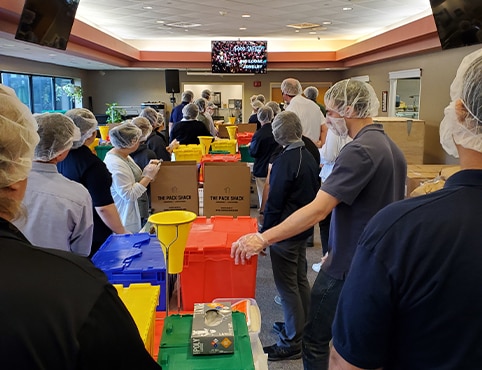A group of Benjamin Moore employee volunteers with colored plastic containers watching an instructional video on how to safely pack food kits. A group of Benjamin Moore employee volunteers with colored plastic containers watching an instructional video on how to safely pack food kits.