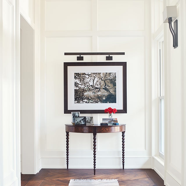 A bright, white-painted hallway with elegant board and batten walls, a piece of framed art, a table, tan striped rug, and dark wood floor. A bright, white-painted hallway with elegant board and batten walls, a piece of framed art, a table, tan striped rug, and dark wood floor.