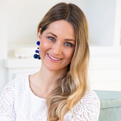 A head shot of a Caucasian woman with wavy blonde hair wearing a white blouse with blue earrings.