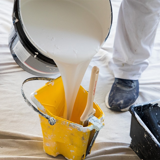 A bucket of white paint being poured into a smaller yellow bucket on top of a white drop cloth. A bucket of white paint being poured into a smaller yellow bucket on top of a white drop cloth.