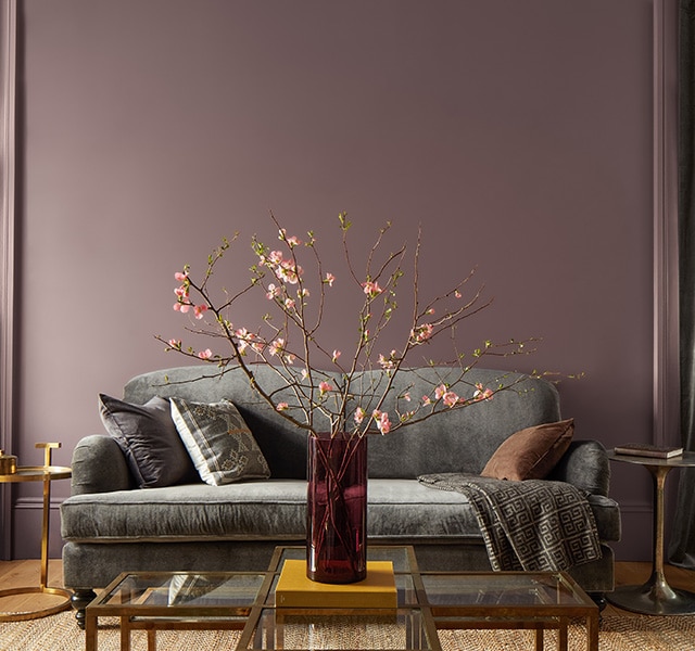 A purple-painted living room features a gray couch, gold side tables, and a glass-topped gold coffee table topped with a red vase filled with pink flowers. A purple-painted living room features a gray couch, gold side tables, and a glass-topped gold coffee table topped with a red vase filled with pink flowers.