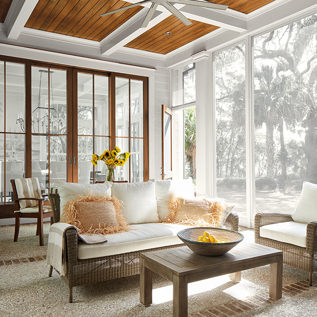 A beautiful screened-in porch with a wood paneled and white beamed ceiling, white trim, a ceiling fan and white cushioned furniture. A beautiful screened-in porch with a wood paneled and white beamed ceiling, white trim, a ceiling fan and white cushioned furniture.