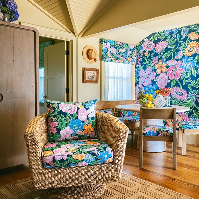 A dark tan painted dining, living room space with rattan and wood furniture, colorful floral wallpaper, and matching floral cushions and pillows. A dark tan painted dining, living room space with rattan and wood furniture, colorful floral wallpaper, and matching floral cushions and pillows.