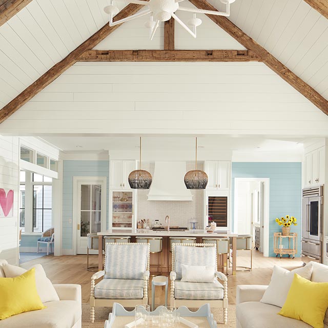 An airy, open white-painted living room and kitchen with shiplap walls and vaulted ceiling, wood beams, blue and white decor, and far back blue walls. An airy, open white-painted living room and kitchen with shiplap walls and vaulted ceiling, wood beams, blue and white decor, and far back blue walls.